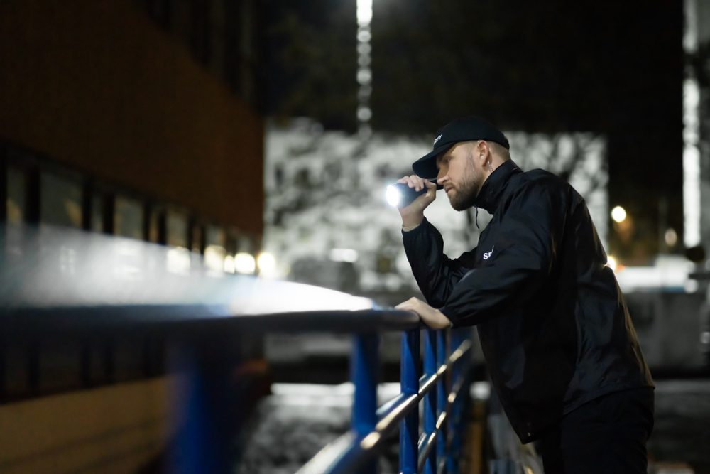 Security,Guard,Walking,Building,Perimeter,With,Flashlight,At,Night
