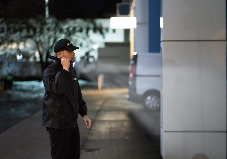 Security,Guard,Walking,Building,Perimeter,With,Flashlight,At,Night