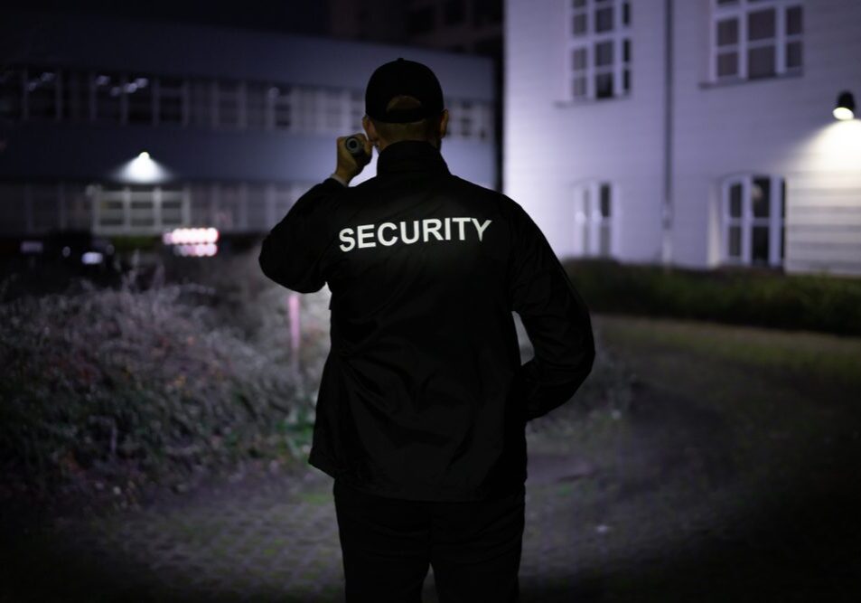 Security,Guard,Walking,Building,Perimeter,With,Flashlight,At,Night