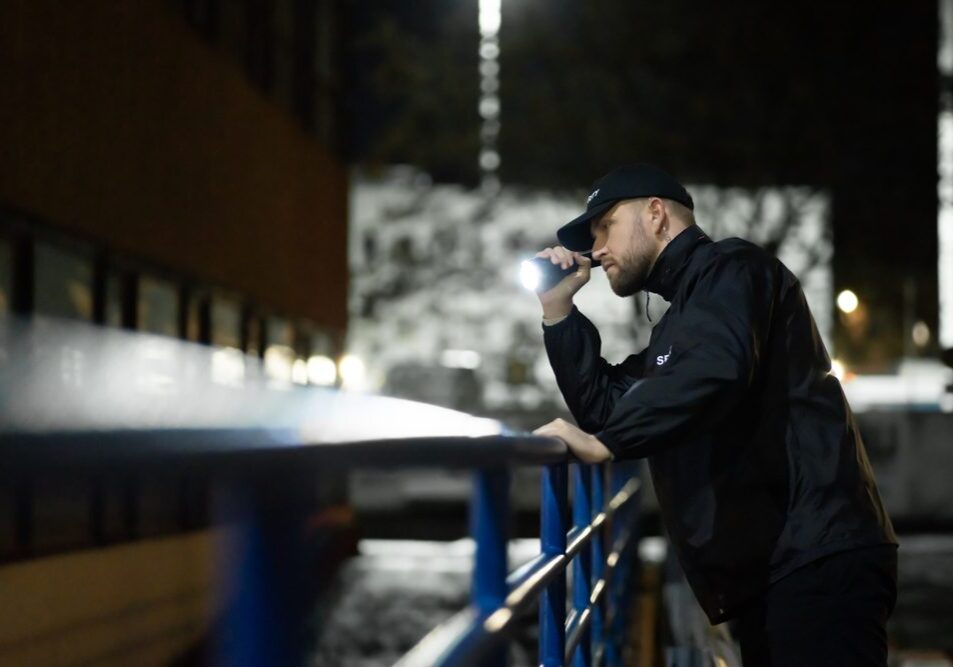 Security,Guard,Walking,Building,Perimeter,With,Flashlight,At,Night